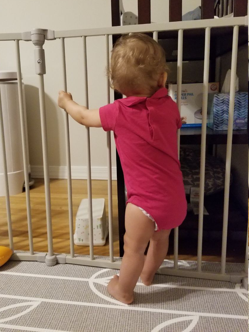 My oldest daughter, aged around 6 months old, facing away from the camera, standing by holding on to the bars of her play pen.